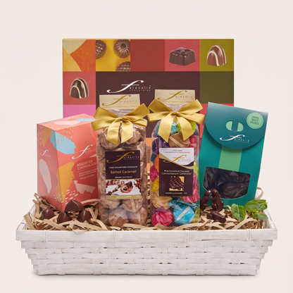 “Fardoulis Celebration Gourmet Chocolate Hamper in a white basket with assorted chocolate boxes, gold-bowed gift bags and dark mint frogs.”
